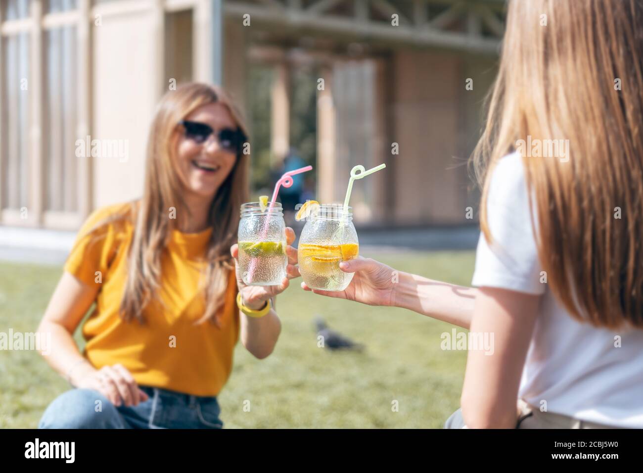 Due belle ragazze nella città soleggiata estate che bevono un cocktail da vasi di vetro. Seduto sull'erba nel parco. Due amici ridenti si rilassano con lemo Foto Stock