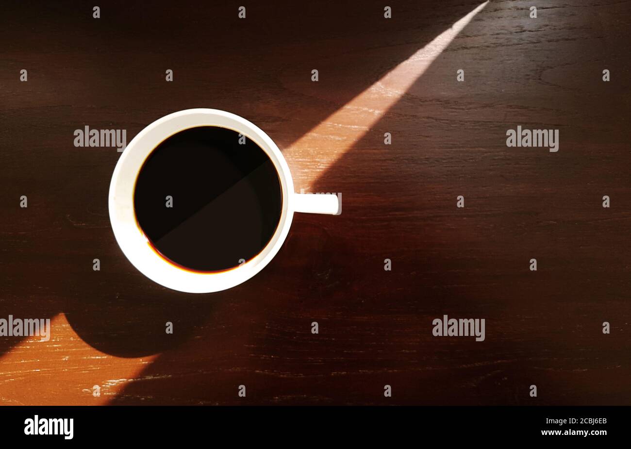 Tazza da caffè in ceramica bianca con caffè espresso nero su sfondo tavolo in legno con luce solare triangolare e ombra dalla finestra, vista dall'alto Foto Stock