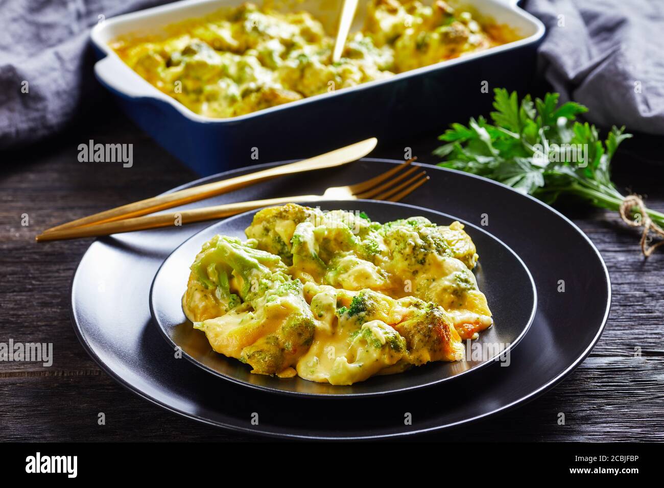 Una porzione di torta di Cheesy Broccoli, casseruola di broccoli su una piastra nera su un tavolo di legno con piatto da forno sullo sfondo, vista orizzontale da abov Foto Stock
