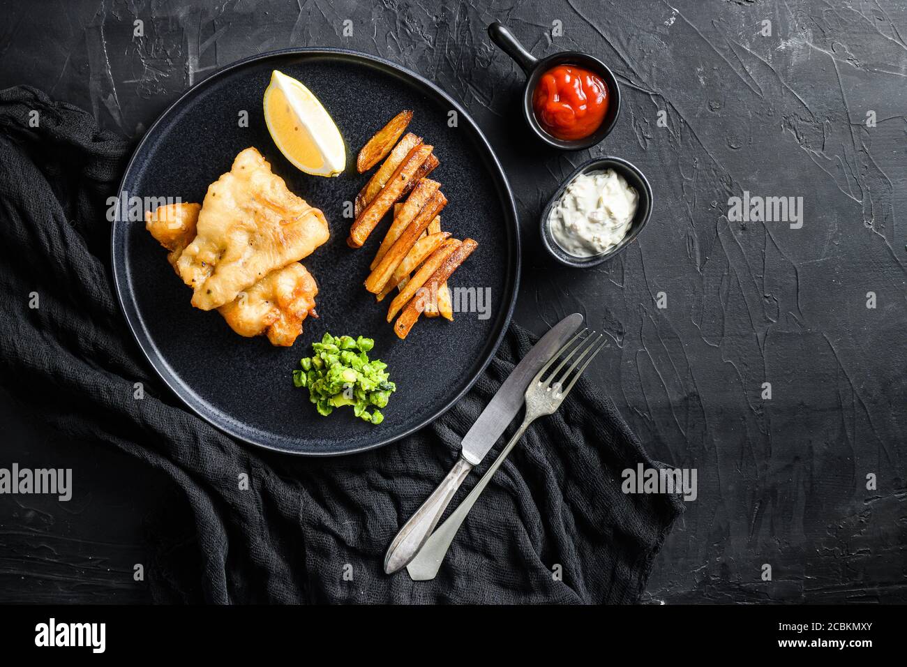Pesce e patatine. Tradizionale piatto britannico di pesce fritto, patatine fritte, piselli minati e salsa tartare e ketchup vista dall'alto, pietra nera Foto Stock