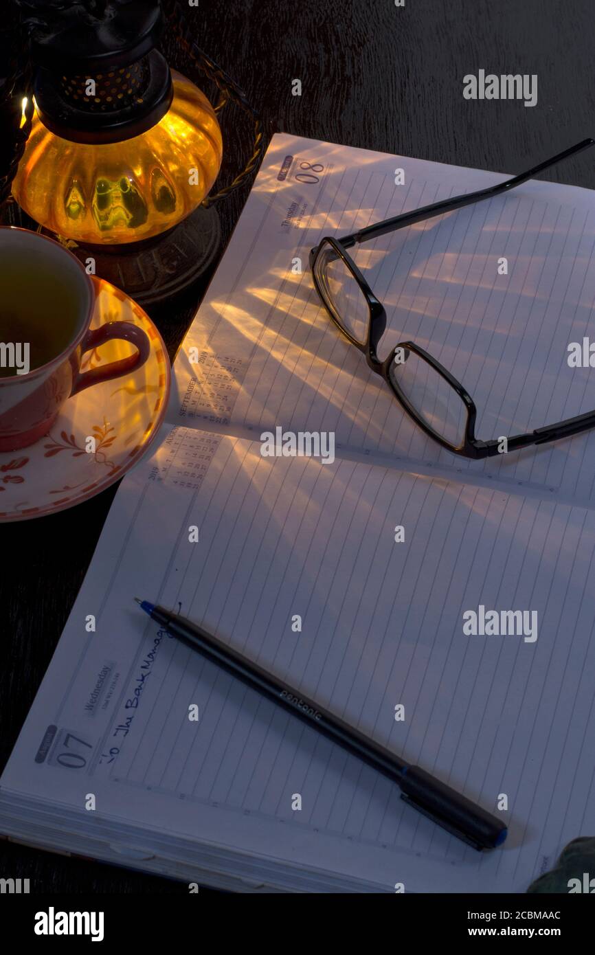Un notebook da ufficio vuoto con occhiali da penna con un lampada ombra e tazza di tè sullo sfondo Foto Stock