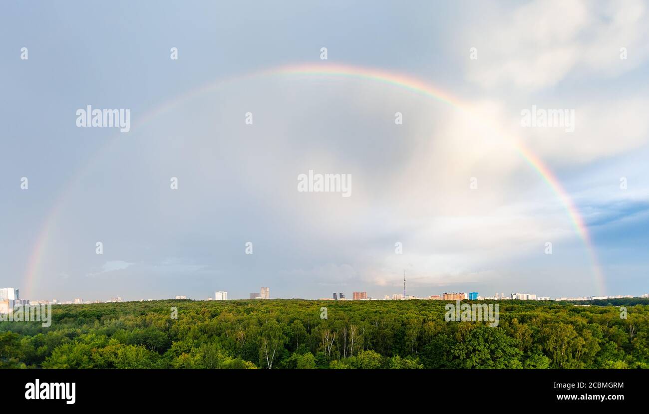 vista panoramica dell'arcobaleno nel cielo blu nuvoloso sul verde foresta e quartiere residenziale in città in estate soleggiata sera Foto Stock