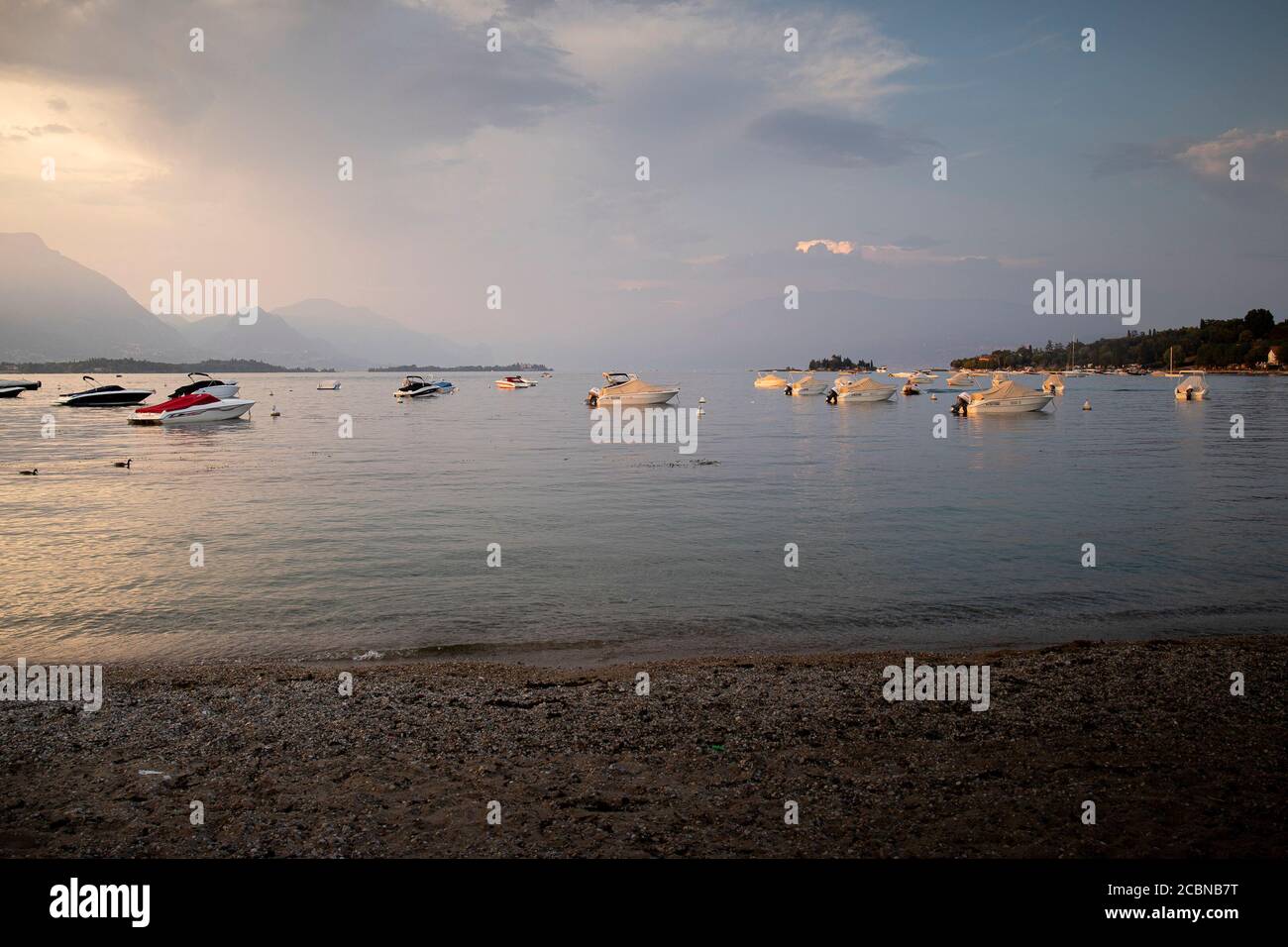 Tramonto tempestoso sulla romantica spiaggia di Manerba del Garda, Lombardia, Italia. Splendida luce dorata su imbarcazioni fisse in acque calme. Orizzontale. Foto Stock
