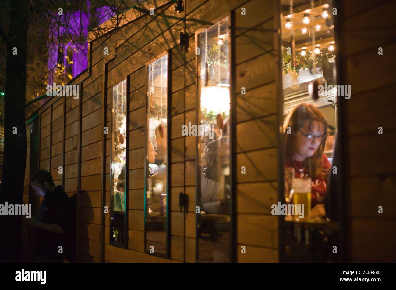 Una donna all'interno di un bar di cabina sul suo telefono, un uomo all'esterno che guarda il suo telefono, mercatino di Natale, feste, birra, notte fuori, fotografia di strada Foto Stock