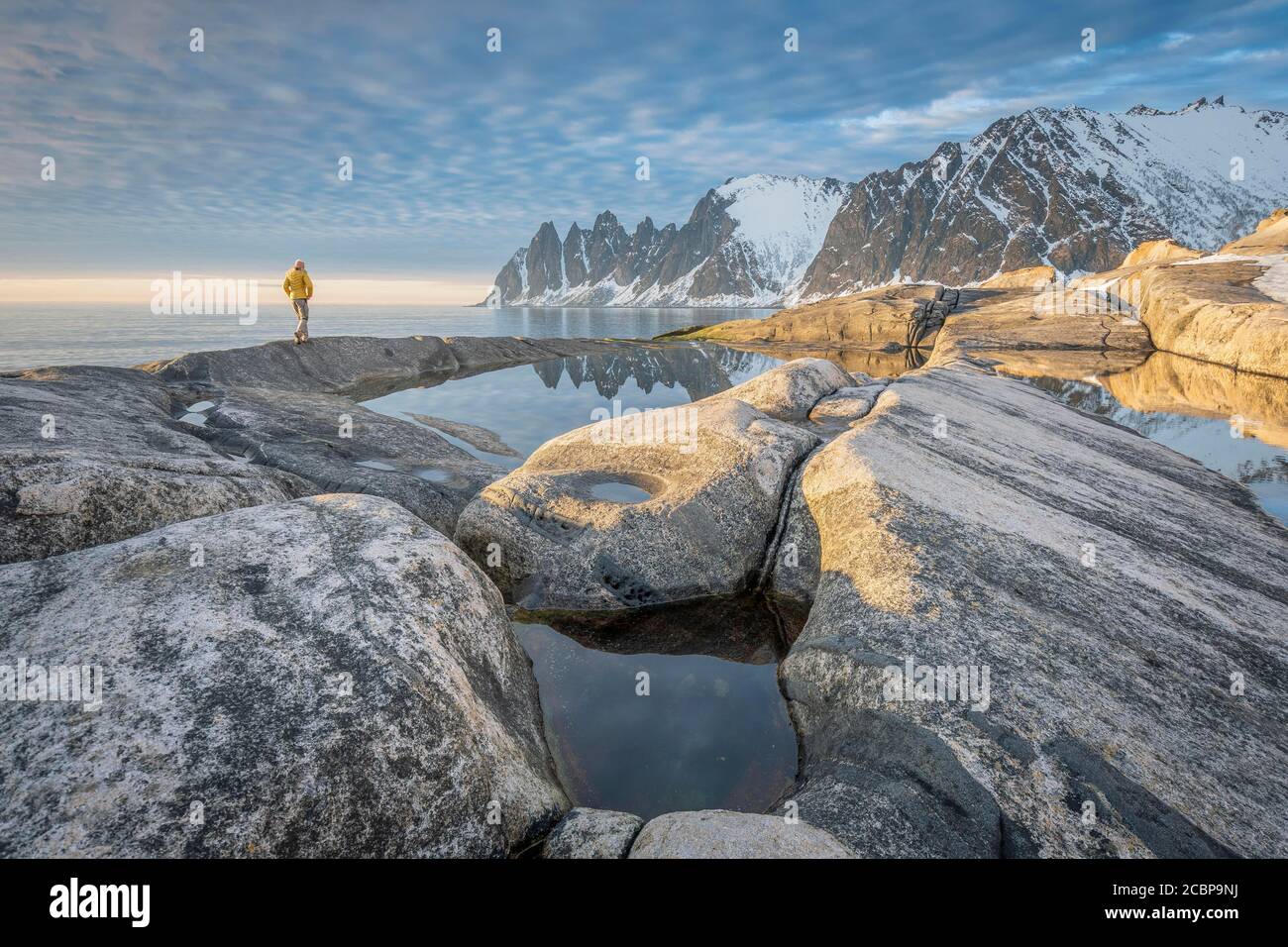 Persona con giacca gialla passeggiate lungo la spiaggia rocciosa di Toggeneset, riflesso delle cime rocciose innevate denti Devils, denti del diavolo Foto Stock