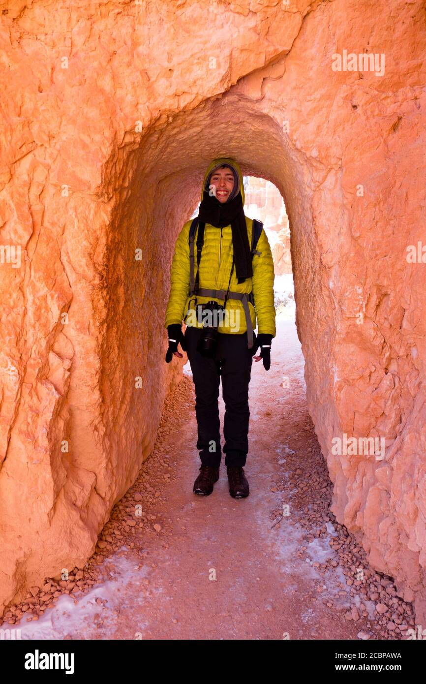 Giovane uomo in abiti invernali con macchina fotografica, sentiero escursionistico attraverso tunnel stretto, escursionista, inverno, Queens Garden Trail, Bryce Canyon National Park, Utah, USA Foto Stock