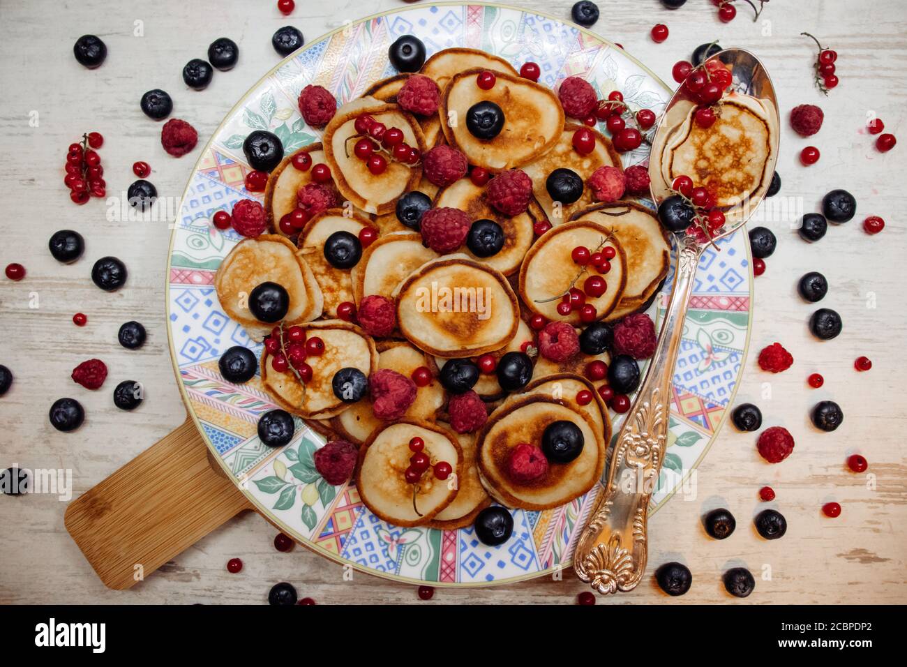 Mini frittelle con frutti di bosco su un piatto a motivi geometrici con cucchiaio d'argento in stile rustico. Colazione tradizionale su legno bianco Foto Stock