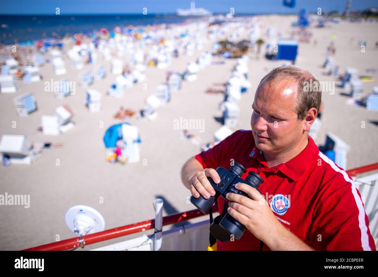 Rostock, Germania. 12 agosto 2020. Il leader dell'orologio Lukas Knaup osserva dalla torre principale dei bagnini sulla spiaggia di Warnemünde con i binocoli nuotatori nel Mar Baltico. Attualmente i bagnini della Croce Rossa tedesca sono sulle torri di guardia ogni giorno e garantiscono le operazioni di balneazione alle spiagge lungo la costa del Mar Baltico. Credit: Jens Büttner/dpa-Zentralbild/dpa/Alamy Live News Foto Stock