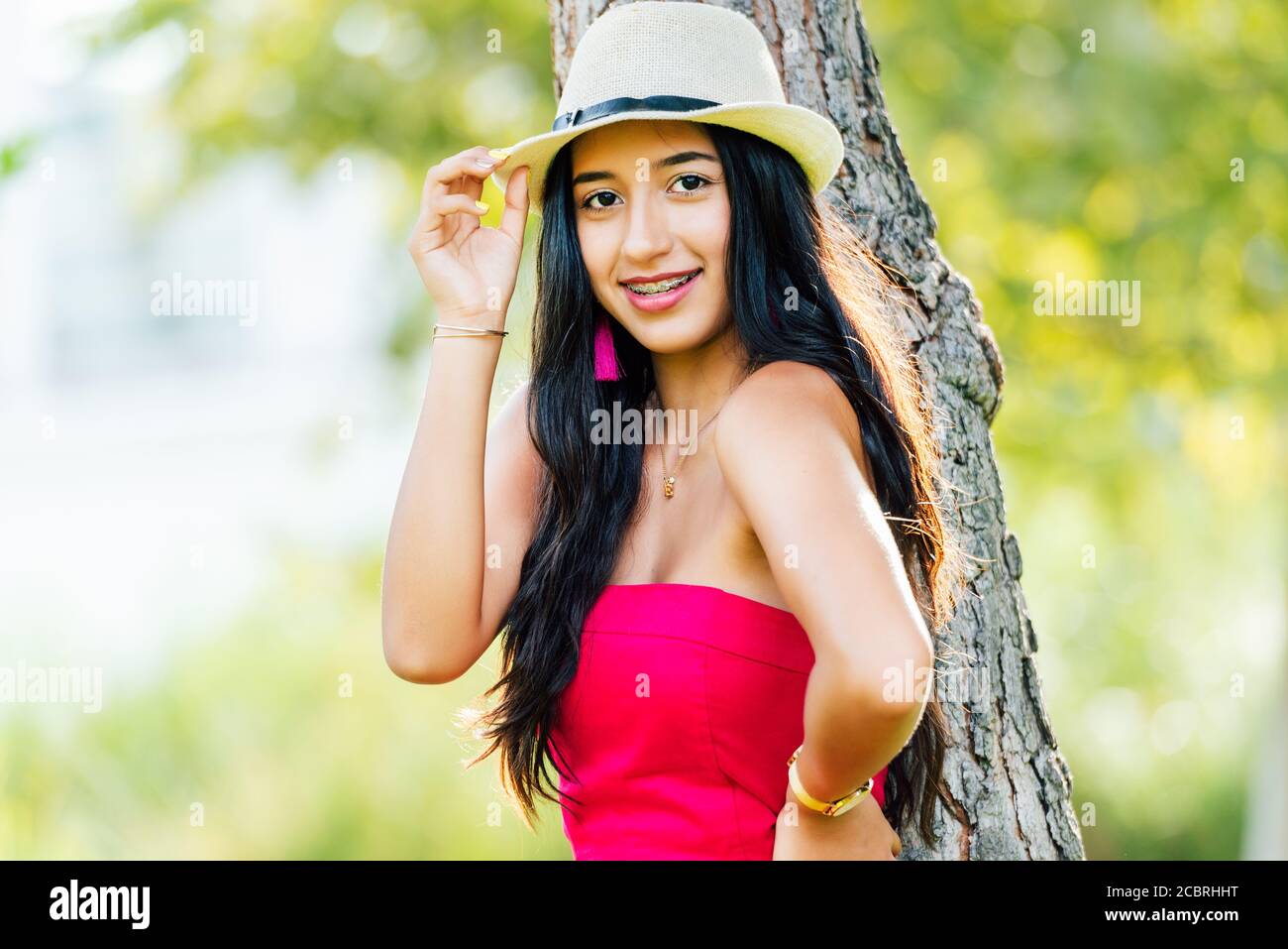 Giovane bruna Latina con lunghi capelli neri e occhi neri, indossando un abito rosso e un cappello leggero, sorridendo e guardando la macchina fotografica appoggiata sul tronco o Foto Stock