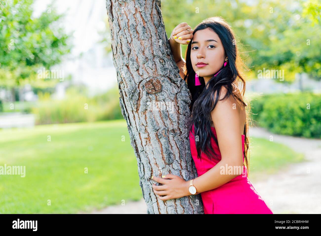 Giovane ragazza Latina bruna con lunghi capelli neri e occhi neri, indossando un vestito rosso, guardando la macchina fotografica appoggiata su un tronco di albero. Copia spazio Foto Stock