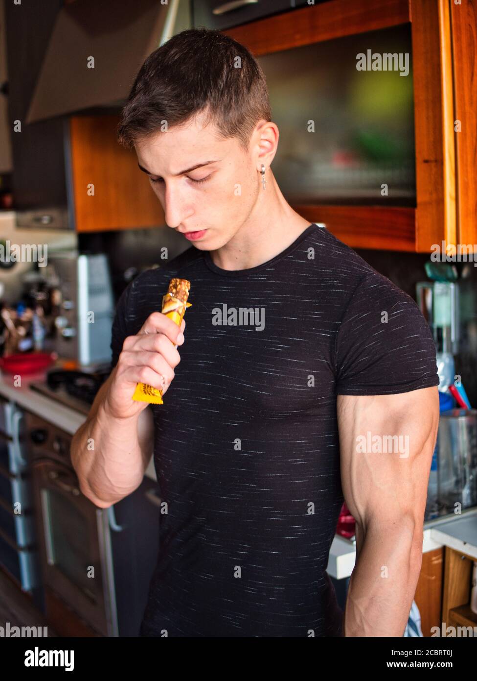 Giovane uomo muscoloso che mangia la barra dei cereali, guardando la macchina fotografica in piedi, Foto Stock