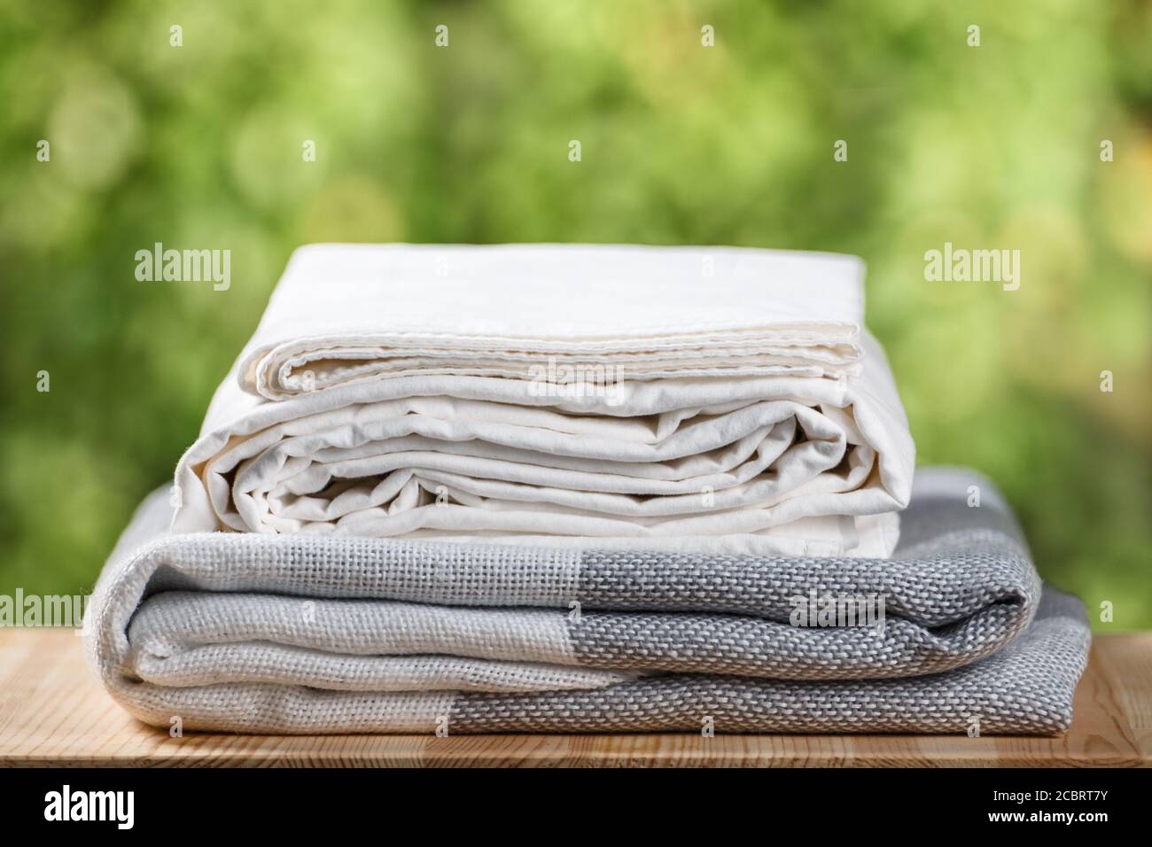 Una pila di biancheria da letto piegata. Foglio di tessuto bianco bianco bianco bianco grigio monocromo su sfondo di fogliame sfocato. Foto Stock