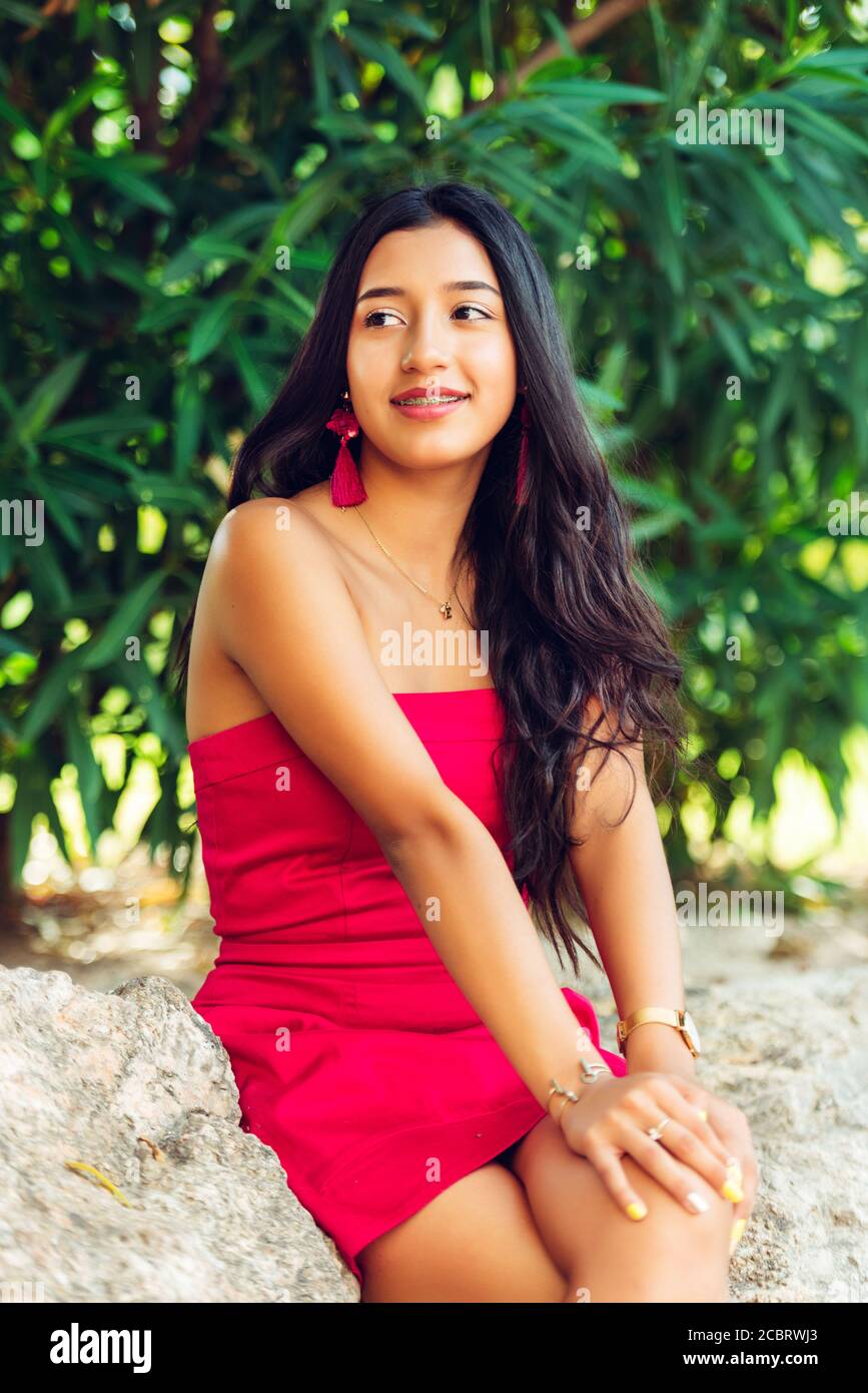 Giovane ragazza latina sorridente con lunghi capelli neri e vestito rosso, in una zona di vegetazione e rocce. Foto Stock