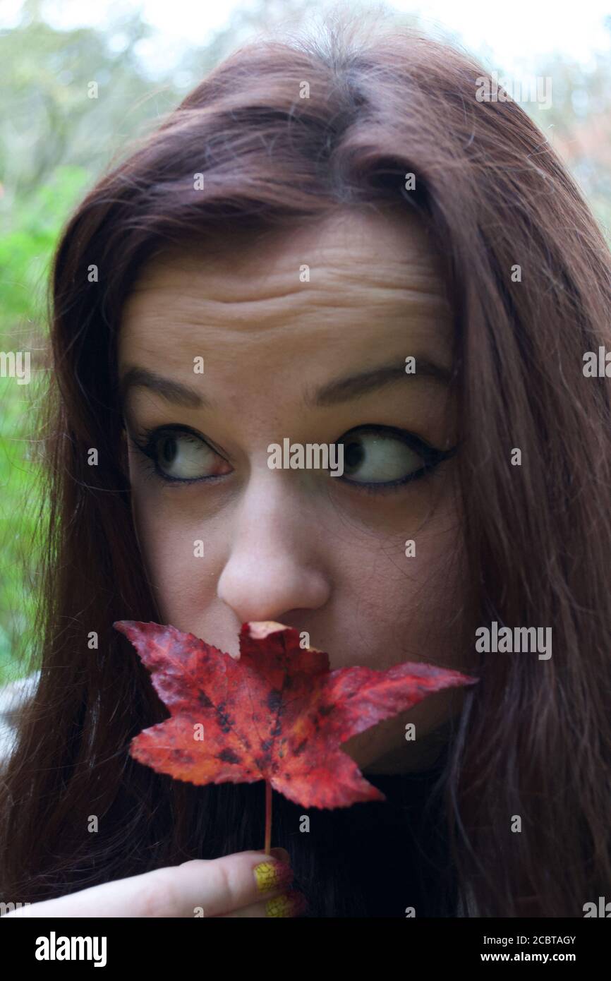 Una giovane donna con occhio pesante compongono e lungo capelli brunette che tengono una foglia rossa caduta acer a lei labbra Foto Stock