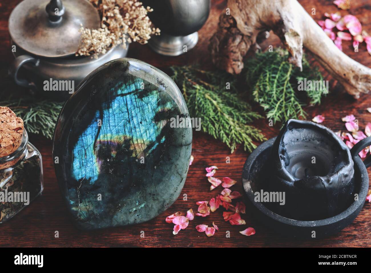 Cristallo di Labradorite su tavolo di legno scuro con vari oggetti naturali come sempreverdi essiccati, piante, fiori, erbe sullo sfondo Foto Stock
