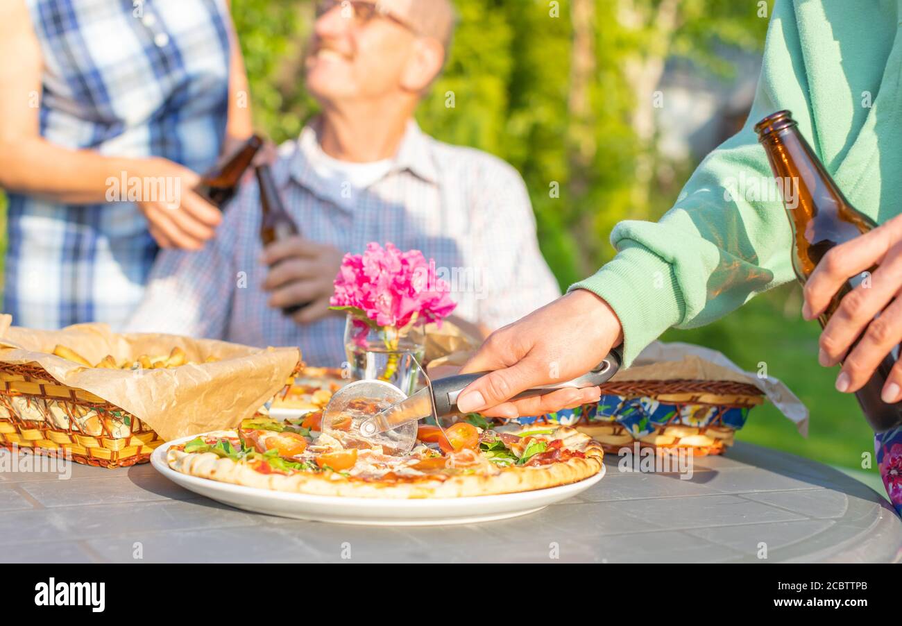 Immagine ritagliata di donna che taglia la pizza durante la festa di famiglia tostatura di una coppia anziana con birra sullo sfondo Foto Stock