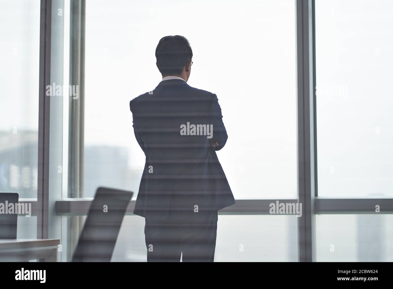 vista posteriore di un uomo d'affari asiatico in piedi davanti di finestre d'ufficio che pensano braccia incrociate Foto Stock