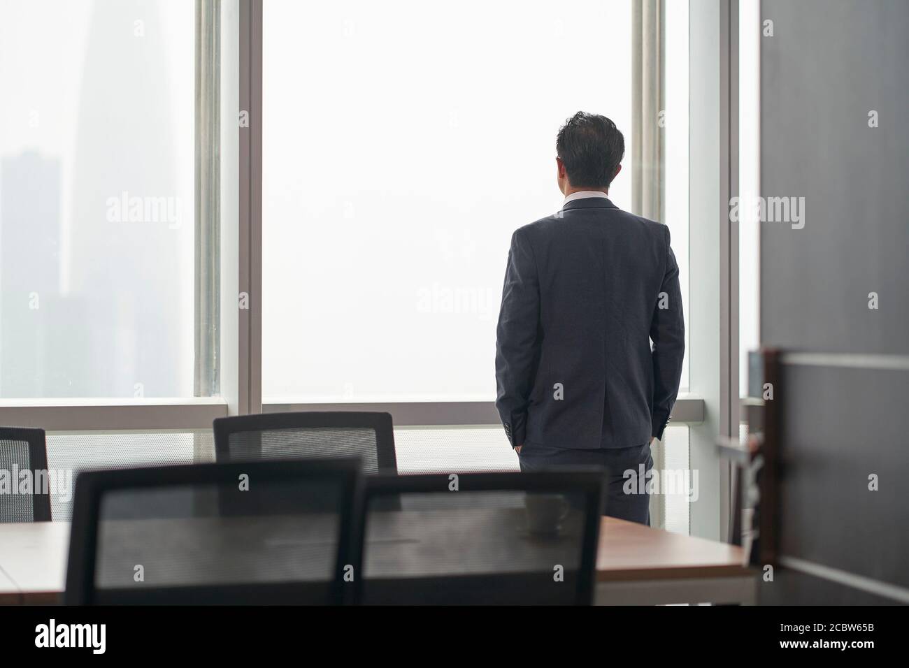 vista posteriore di un uomo d'affari asiatico in piedi davanti della finestra di ufficio che pensa le mani in tasche Foto Stock