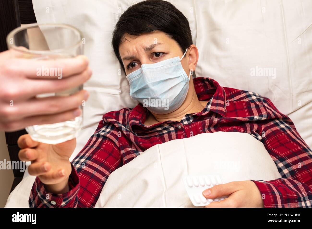 Donna malata in maschera medica che si trova sul letto è dato un bicchiere d'acqua per prendere pillole. La cura familiare per il concetto di malato Foto Stock
