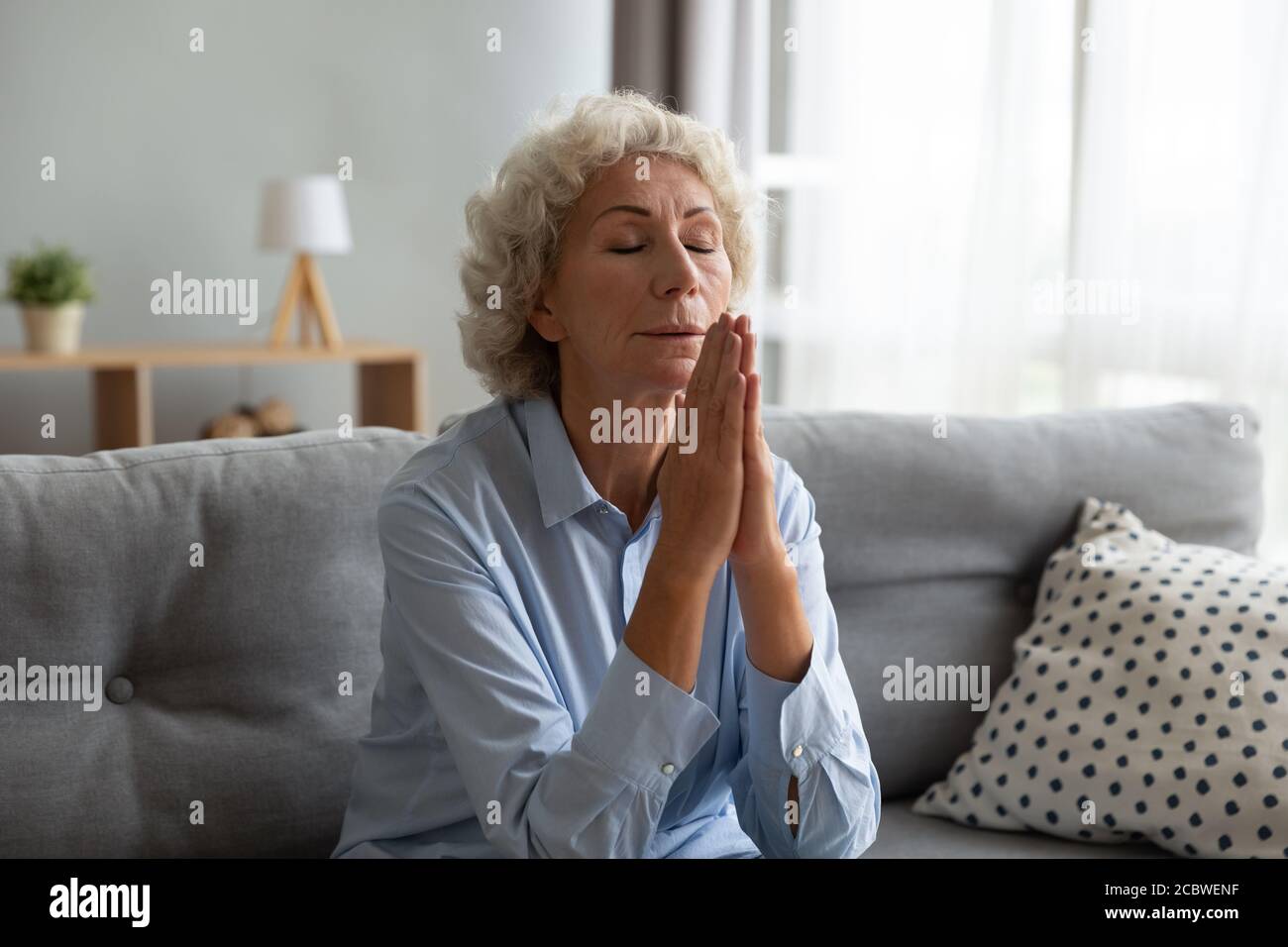 Fedele anziano maturo pregando Dio, seduto sul divano. Foto Stock