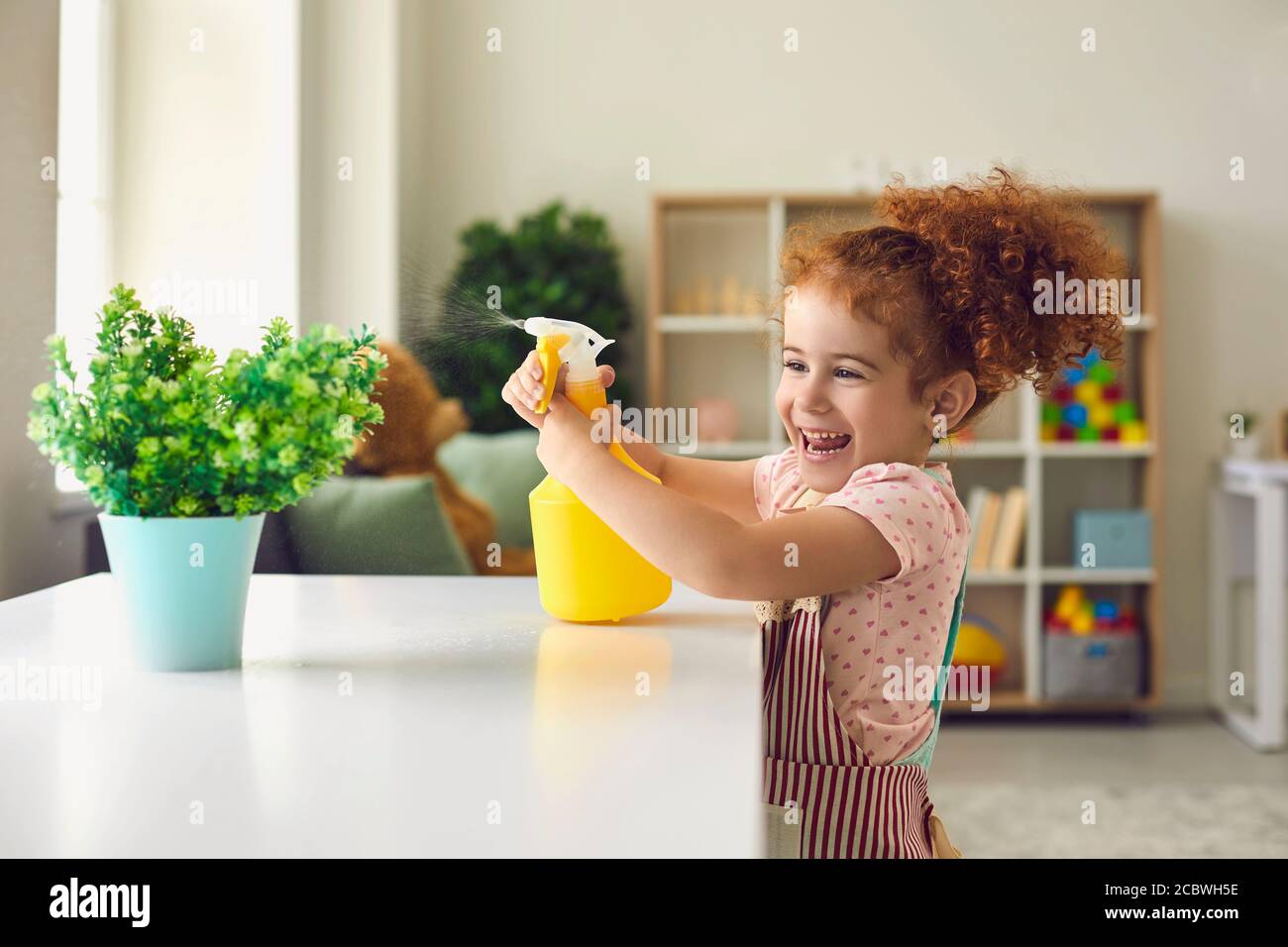 Bambina spruzzando acqua su pianta domestica a casa. Carino bambino che aiuta con i doveri della famiglia. Allegro bambino annaffiando fiore Foto Stock