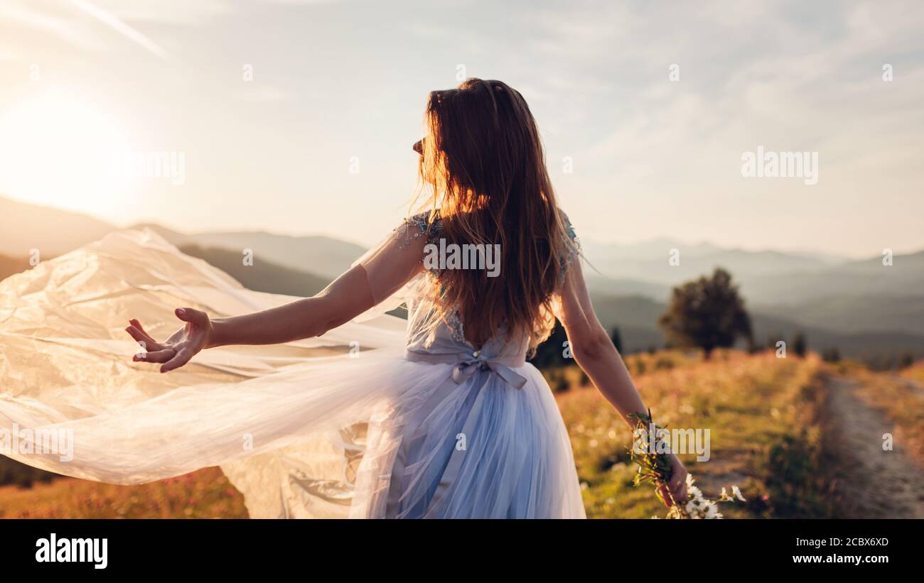 Bella sposa danzare in abito blu matrimonio in montagna al tramonto. Donna lancia il velo che tiene fiori. Foto Stock