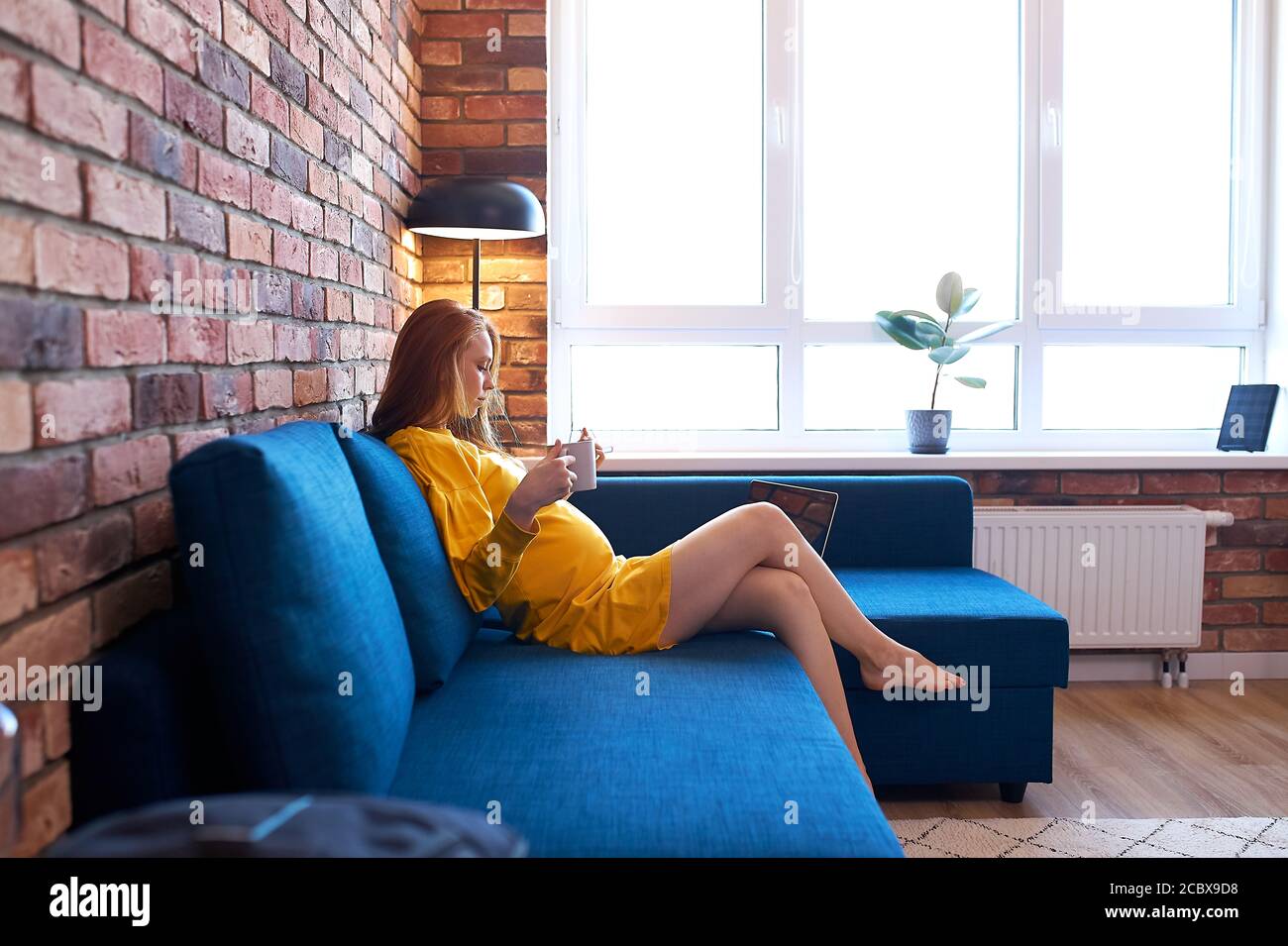vista laterale su attraente donna incinta rosso bere tè a casa, donna in abito giallo si siede sul divano, hanno riposo Foto Stock