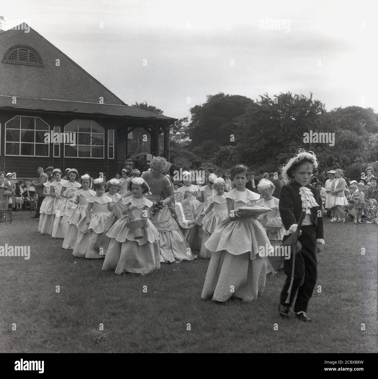 Anni '1950, storico, un giovane ragazzo vestito con un costume da reggenza con merletto cravat che guida la "Rose Queen" e il suo entourage di ragazze, mentre camminano attraverso il parco locale, Farnworth, Lancashire, Inghilterra, Regno Unito. Risalente agli anni '1880, il festival annuale della Regina delle Rose si teneva tradizionalmente nel mese di giugno, diventando un evento annuale in molte città e villaggi in tutto il Regno Unito, specialmente nella contea del Lancashire, nota come contea della Rosa Rossa, dopo le guerre delle rose (1455-87). Foto Stock