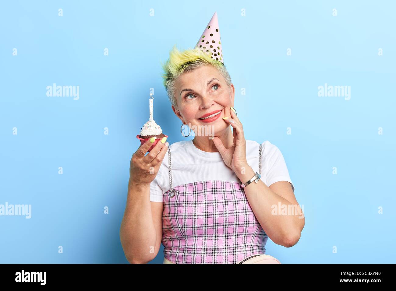 Matura bella donna che tiene una torta di compleanno, toccando la guancia, guardando in su, sognando di amore, felicità. Isolato su sfondo blu Foto Stock