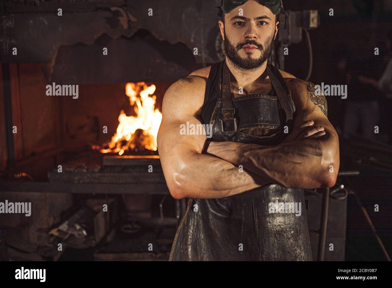 Ritratto di un giovane metalworker serio e sicuro in un grembiule nero che guarda la macchina fotografica, lavora in officina Foto Stock