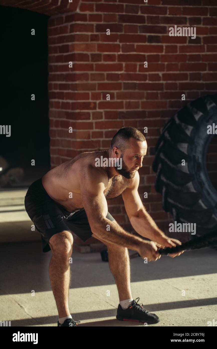 Potente muscolare determinato uomo con formazione di corda in allenamento funzionale in palestra esterna con pareti in mattoni, rafforzare i muscoli delle spalle, w Foto Stock