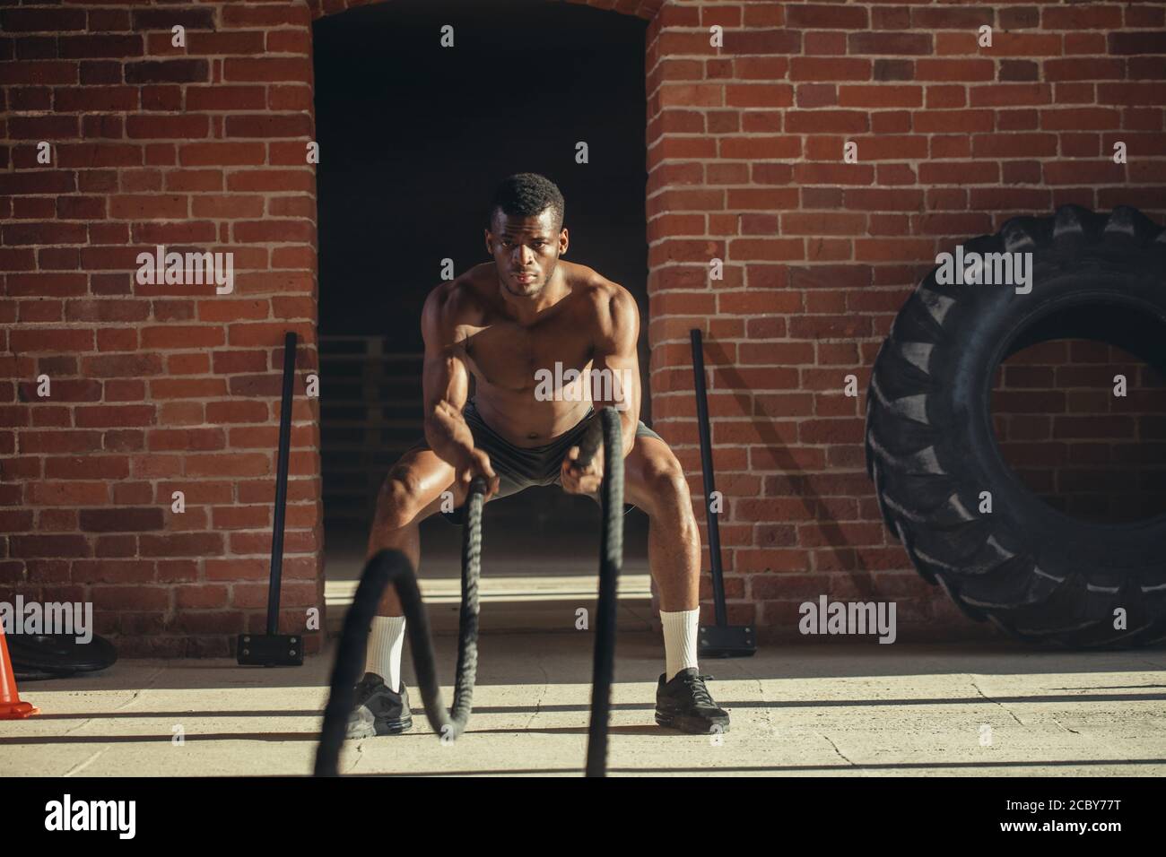 Potente muscolare determinato uomo africano con formazione di corda in allenamento funzionale in palestra esterna con pareti in mattoni, rafforzare i muscoli del shou Foto Stock