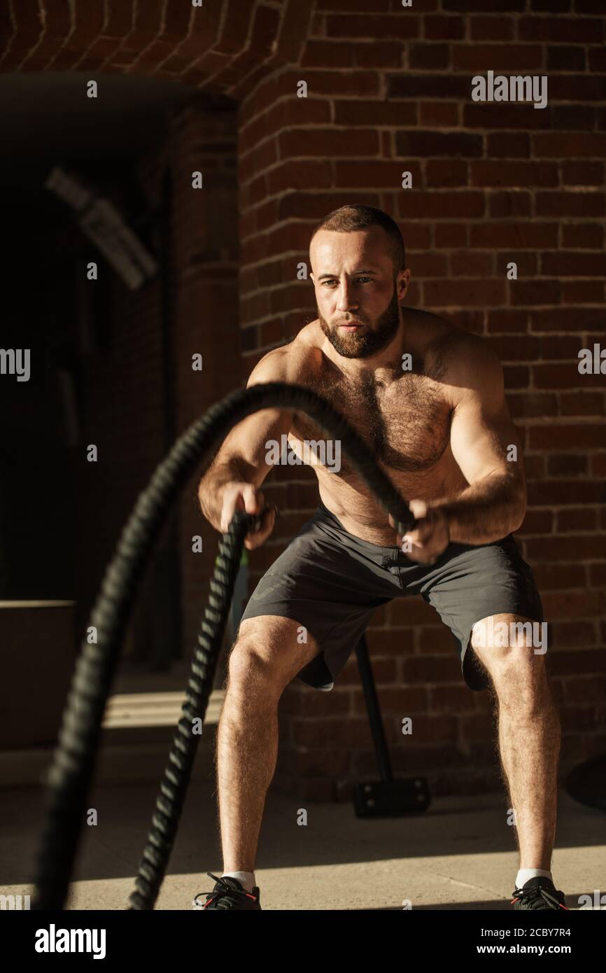 Potente muscolare determinato uomo con formazione di corda in allenamento funzionale in palestra esterna con pareti in mattoni, rafforzare i muscoli delle spalle, w Foto Stock