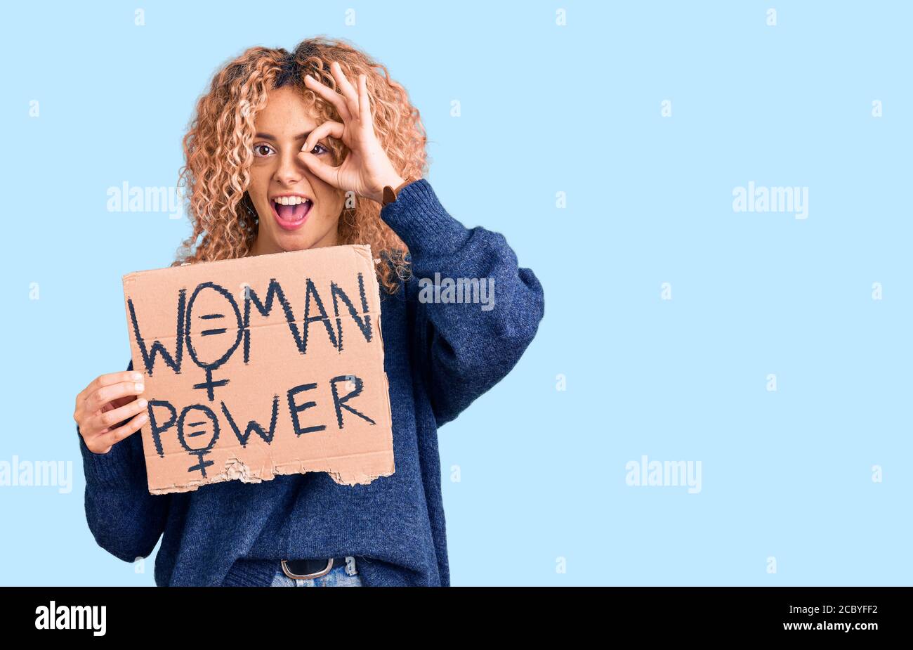 Giovane donna bionda con i capelli ricci che tengono abbiamo bisogno di un cambia banner sorridente felice facendo ok segno con mano sopra occhio che guarda attraverso le dita Foto Stock