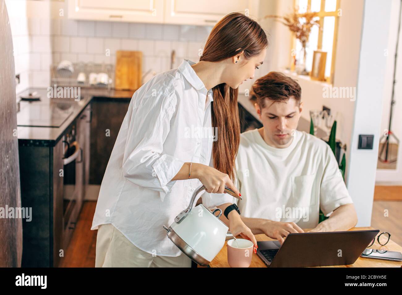 donna caucasica gentile tratta il suo marito con tè gustoso mentre sta lavorando da casa, usando il laptop. moglie misericordiosa ama il marito, lo sostiene. freelanc Foto Stock