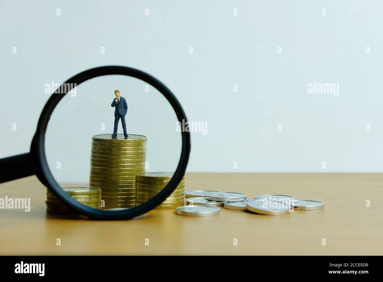 Concetto di business di persone in miniatura - un uomo d'affari in piedi su pila di monete con lente di ingrandimento, alla ricerca di soluzione finanziaria / strategia Foto Stock