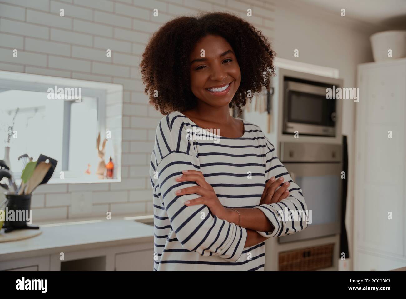 Ritratto di giovane donna africana allegra in piedi in cucina moderna con le braccia ripiegate che guardano la fotocamera Foto Stock