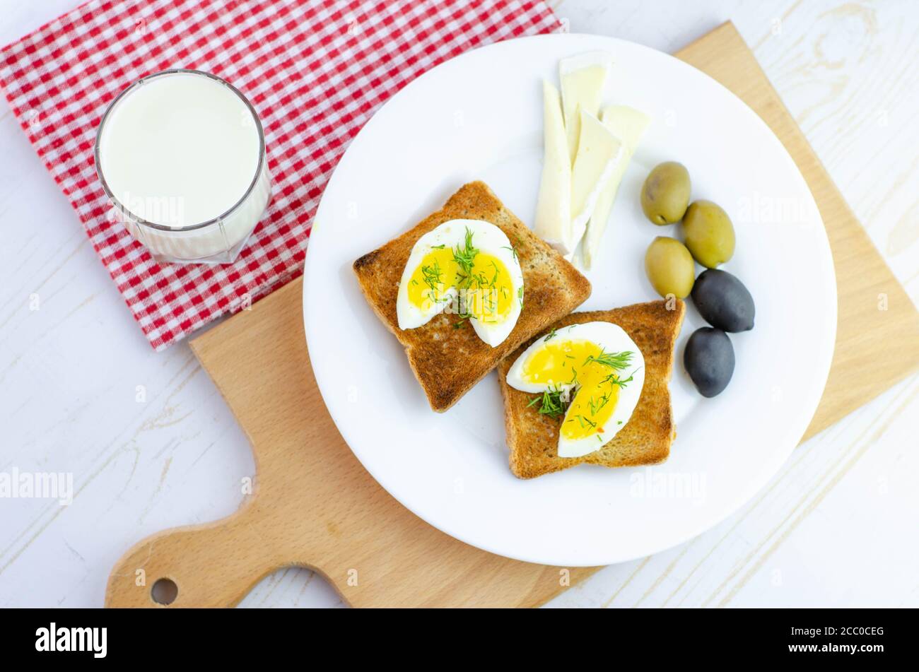 Toast fresco con uova, olive e formaggio di soia per colazione e un bicchiere di latte su sfondo di legno bianco con tagliere di legno e asciugamano a scacchi Foto Stock