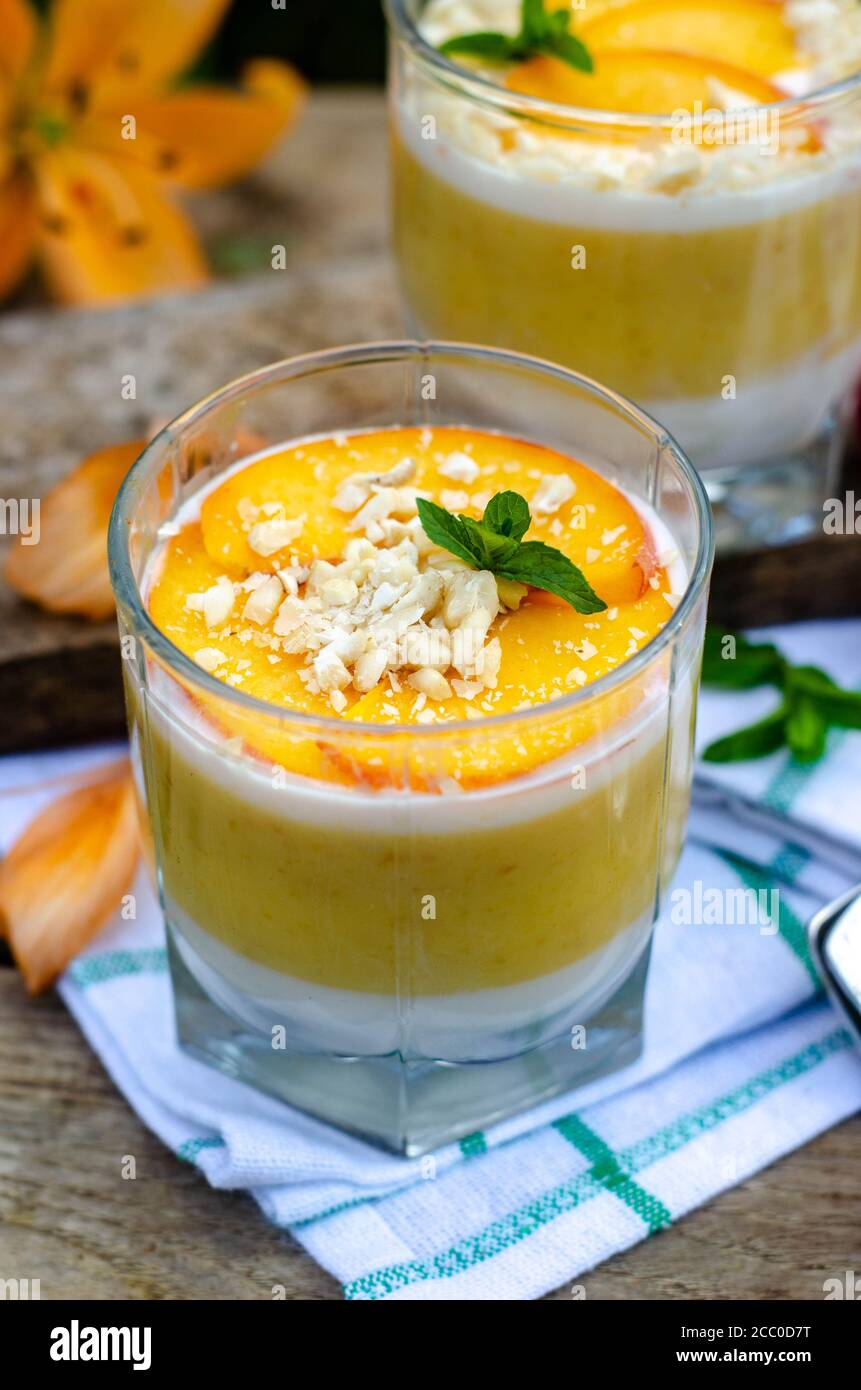 Due galassine di panna cotta di pesca a strati bianchi e arancioni. Servita all'aperto su sfondo di legno con menta e ciliegia. Foto Stock