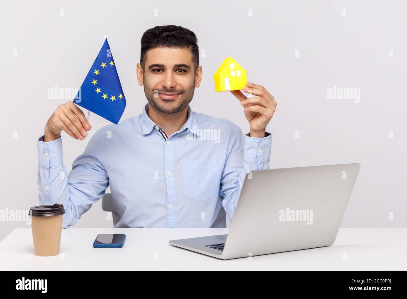 Giovane positivo seduto in ufficio, con casa di carta e bandiera dell'Unione europea, sorridendo alla macchina fotografica, pubblicità di acquisto di beni immobili Foto Stock