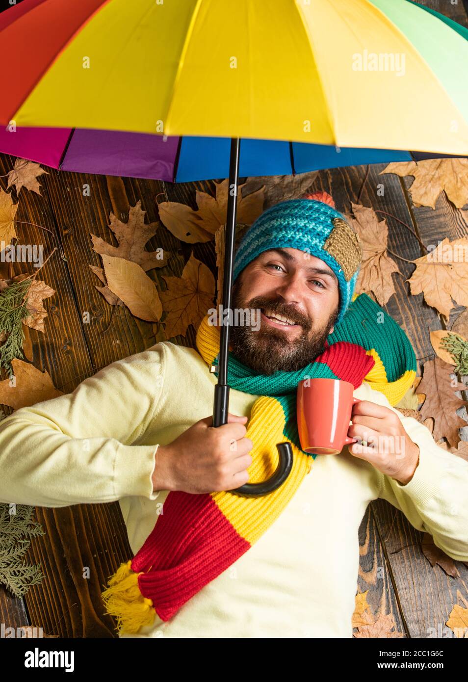 Uomo allegro che beve tè caldo o caffè. Sconti autunnali sui vestiti. Uomo  con ombrello color arcobaleno. Gli piace la pioggia, le previsioni meteo  promettono la pioggia. Uomo bearded che tiene l'ombrello