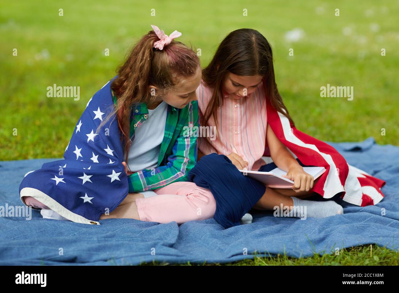 Ritratto a lunghezza intera di due ragazze carine coperte da bandiera americana seduta su coperta da picnic nel parco e libro di lettura, spazio di copia Foto Stock