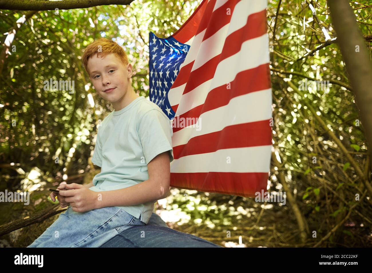 Ritratto di un ragazzo adolescente dai capelli rossi che guarda la macchina fotografica all'aperto mentre si nascondono sotto rami di grande albero in foresta o. Giocare nel cortile posteriore con bandiera americana in background Foto Stock