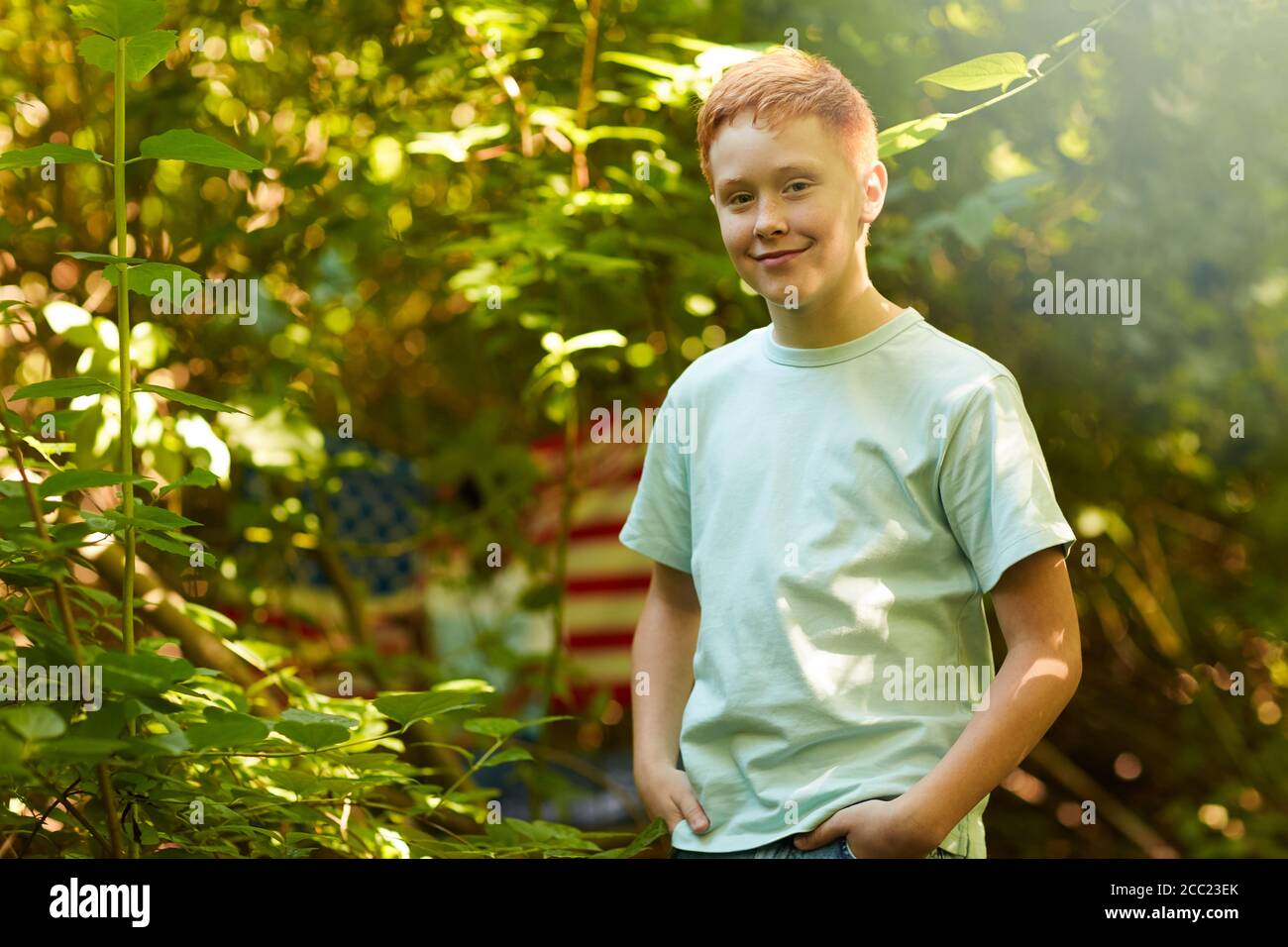 Vita in su ritratto di ragazzo adolescente con capelli rossi sorridente alla macchina fotografica mentre si sta in piedi all'aperto nella foresta o cortile posteriore, copia spazio Foto Stock