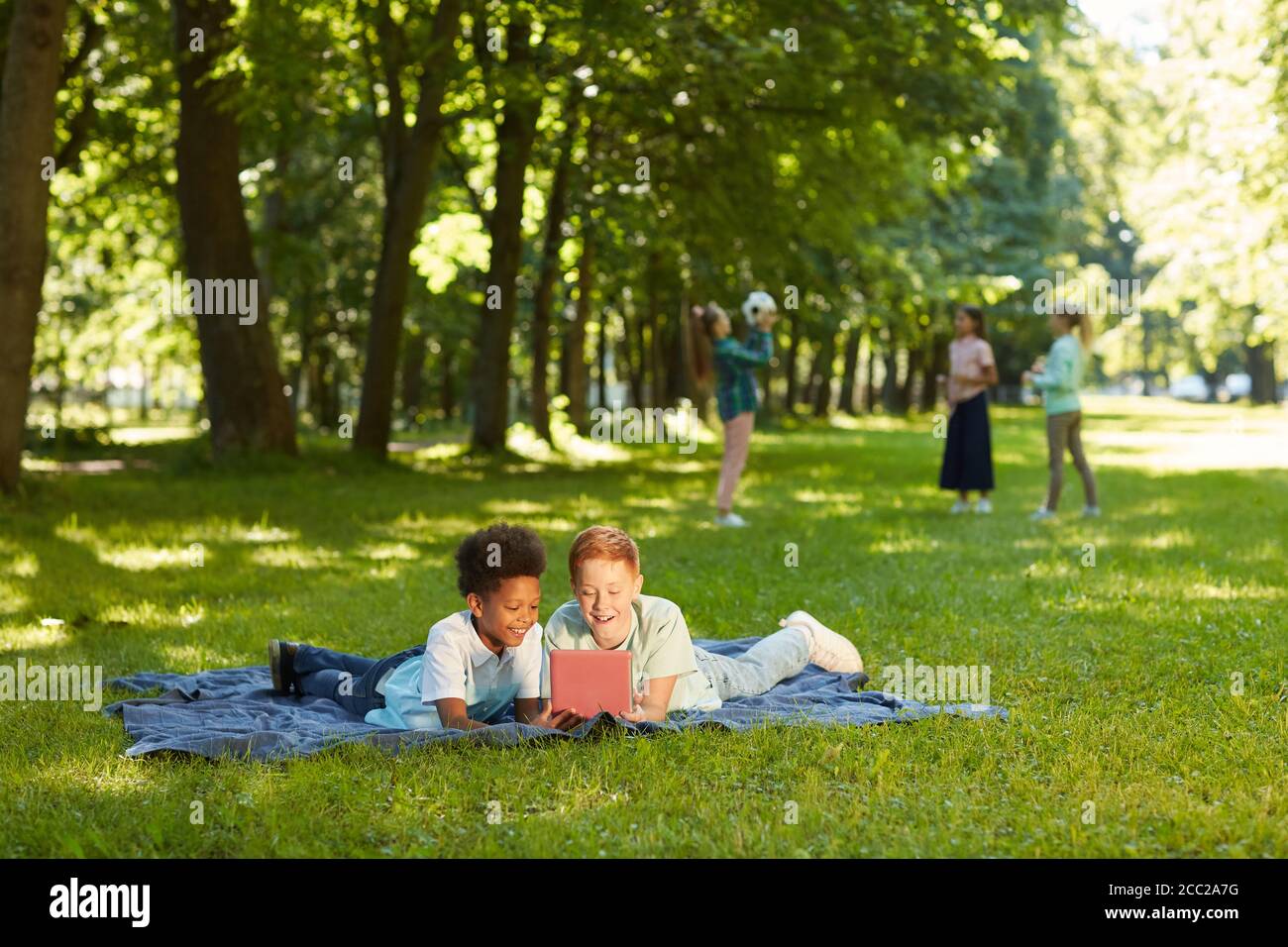 Ritratto grandangolare di due allegri ragazzi che usano il tablet digitale mentre giacciono su erba verde in un parco all'aperto illuminato dalla luce del sole, copia spazio Foto Stock