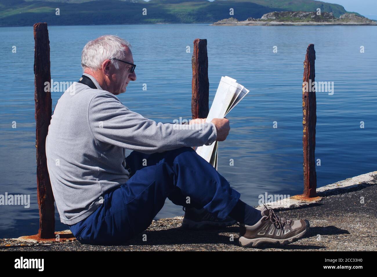 Un uomo più anziano di 60 anni seduto su un molo disusato poggiato su un palo arrugginito guardando la sua mappa contro un mare blu profondo, Argyll, Scozia Foto Stock