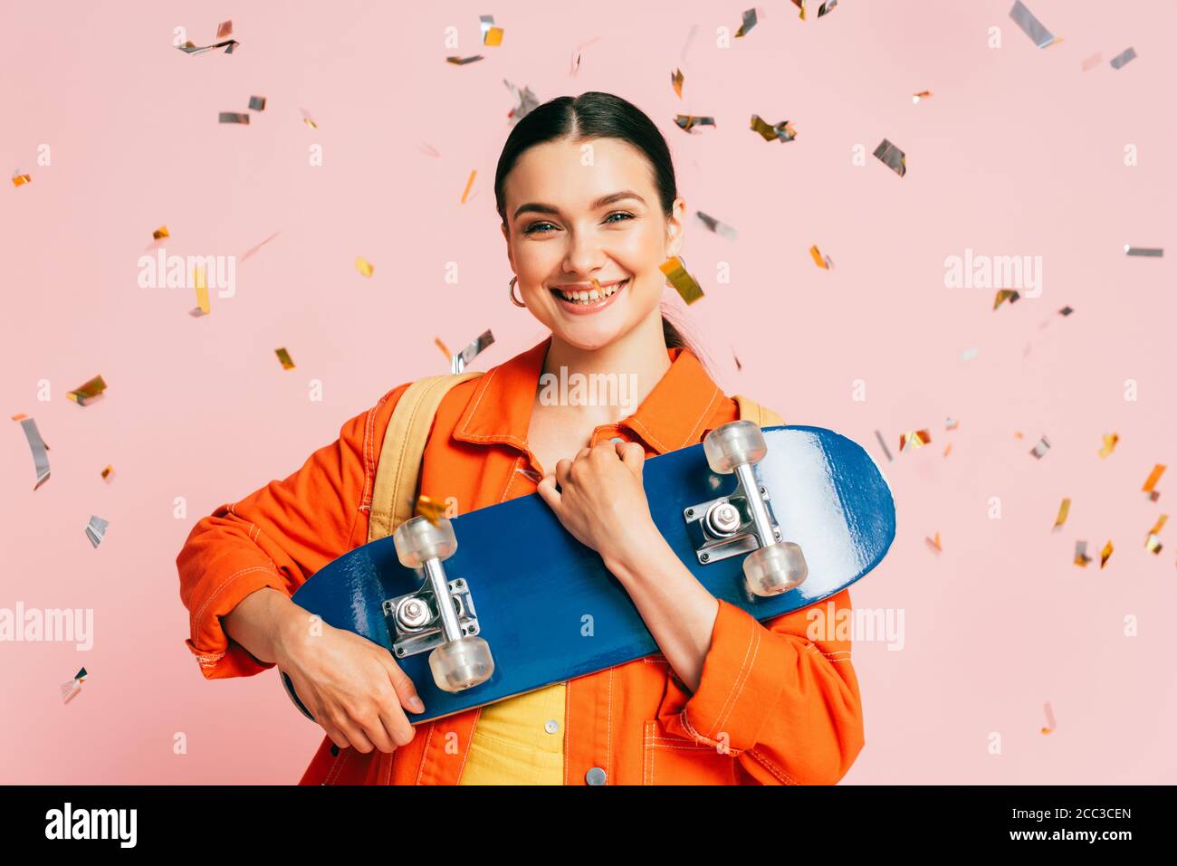 giovane donna bruna in vestito colorato con skateboard sotto caduta confetti isolati su rosa Foto Stock