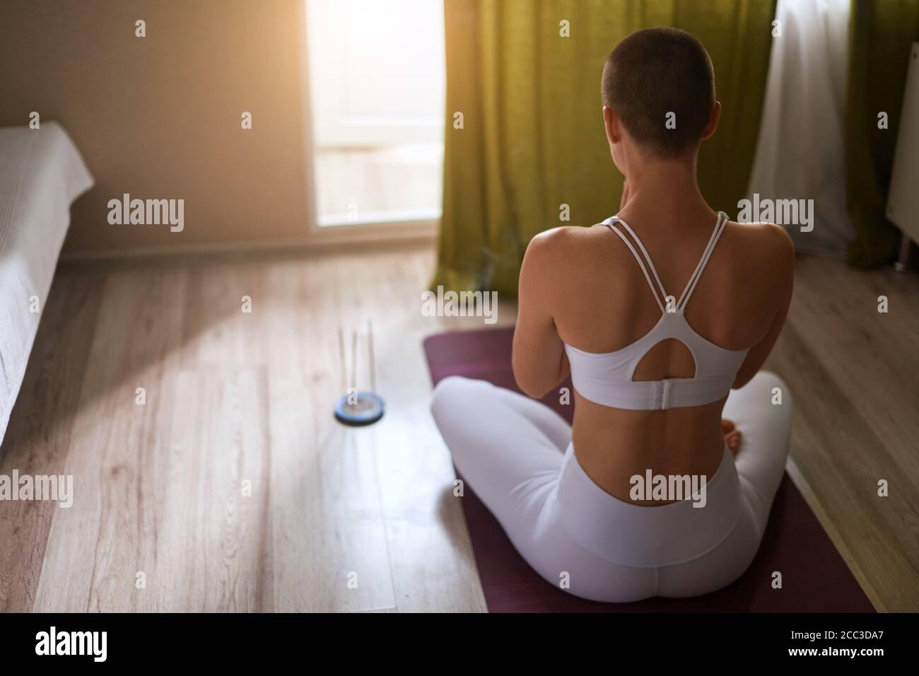 Donna meditando in ambienti chiusi. Attraente e a pelo corto modello caucasici in bianco sportclothes seduta sul tappeto e praticare lo yoga con candele. Pregare Foto Stock