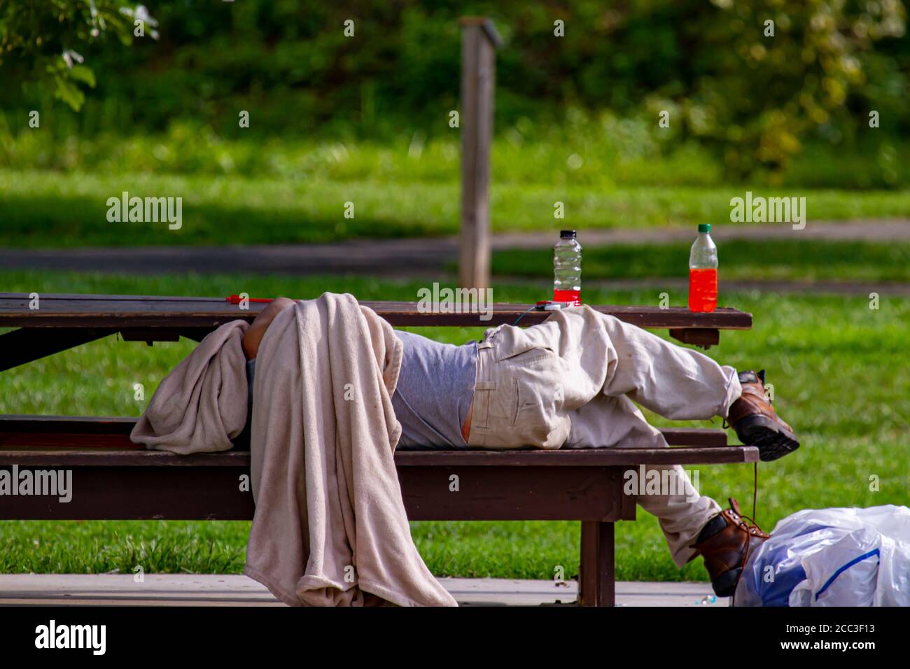 Un uomo caucasico senza casa sta dormendo su un tavolo da picnic in una zona ombreggiata di un parco. Sta coprendo la testa con una coperta sporca. I suoi effetti personali sono dentro Foto Stock