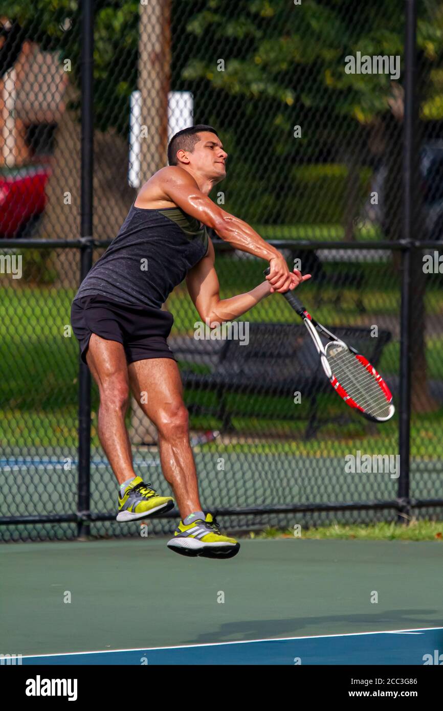 MD, USA 08/14/2020: Un atletico ispanico che indossa una camicia e un pantaloncini senza maniche sudati sta prendendo un servizio durante una partita di tennis in un campo locale a Ba Foto Stock